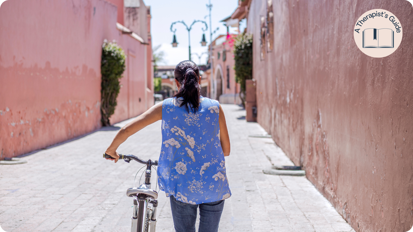brunette woman in blue floral shirt pushing bike down alleyway
